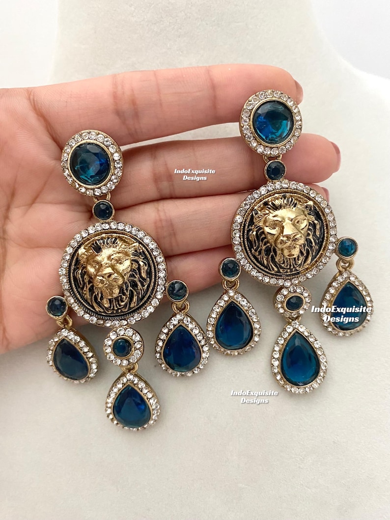 May include: A pair of gold-tone earrings with a lion head design and blue crystal accents. The earrings feature a large round pendant with a lion head in the center, surrounded by smaller blue crystals. The lion head is detailed with a mane and a crown. The earrings are suspended from a smaller blue crystal drop.