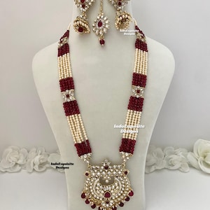 May include: A traditional Indian bridal necklace set featuring a large pendant, red and white beads, and matching earrings. The pendant is decorated with gold and red stones. The set is called "IndoExquisite Designs".
