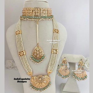 May include: A gold and white pearl necklace set with a choker, long necklace, earrings, and a maang tikka. The necklace features a large, ornate pendant with a floral design and green beads. The earrings are also ornate and feature green beads.