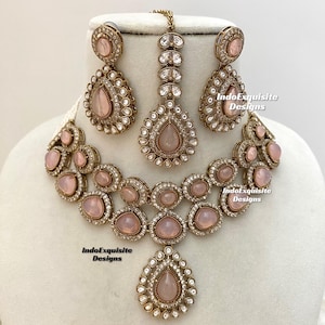 May include: A pink and gold Indian bridal necklace set with a teardrop pendant, matching earrings, and a maang tikka. The set features pink stones and clear rhinestones.