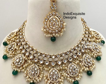 May include: A gold and white Indian bridal necklace set with green beads. The necklace has a layered design with multiple strands of pearls and a large pendant in the center. The set also includes a matching maang tikka, a traditional Indian headpiece.