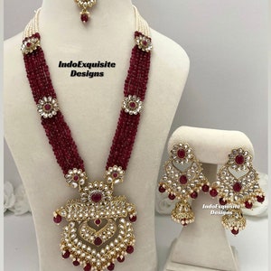 May include: A maroon and gold Indian bridal necklace set with a large pendant, earrings, and a maang tikka. The necklace features multiple strands of maroon beads and pearls, and the pendant is adorned with intricate gold work and red and white stones. The earrings are similarly decorated with gold and stones.