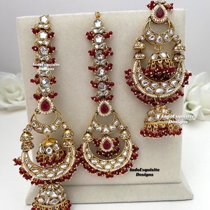Puede incluir: Un conjunto de joyer&iacute;a en tono dorado que incluye dos accesorios para el cabello largos y verticales y un pendiente grande en forma de media luna. Cada pieza est&aacute; adornada con piedras claras, detalles en rojo oscuro y cuentas rojas.