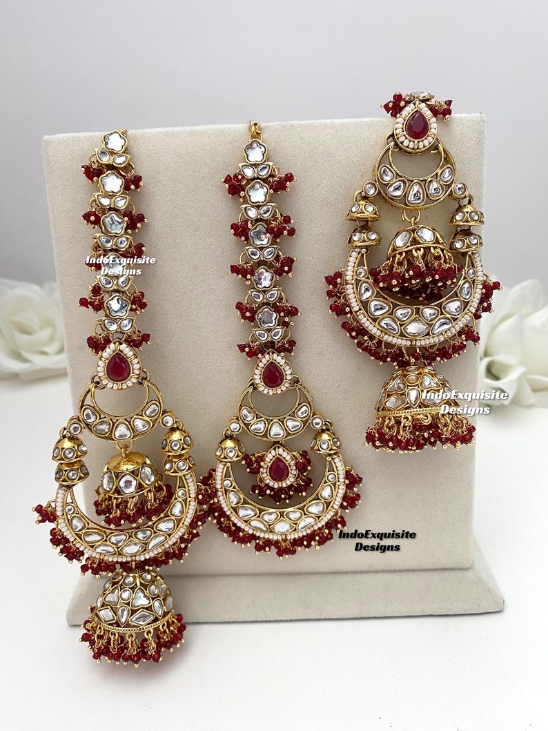 Puede incluir: Un conjunto de joyer&iacute;a en tono dorado que incluye dos accesorios para el cabello largos y verticales y un pendiente grande en forma de media luna. Cada pieza est&aacute; adornada con piedras claras, detalles en rojo oscuro y cuentas rojas.