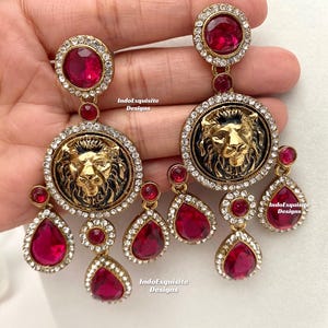 May include: Gold tone earrings with a black enamel lion head design and red crystal accents. The earrings feature a large round pendant with a lion head and smaller teardrop-shaped pendants.