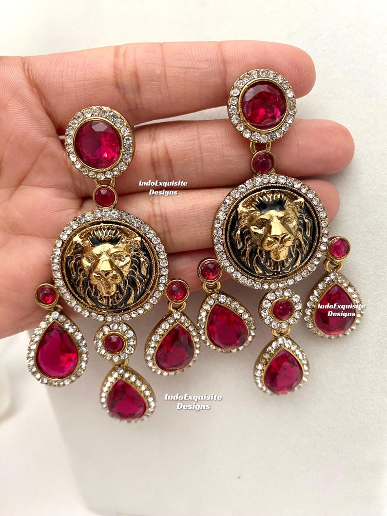 May include: Gold tone earrings with a black enamel lion head design and red crystal accents. The earrings feature a large round pendant with a lion head and smaller teardrop-shaped pendants.