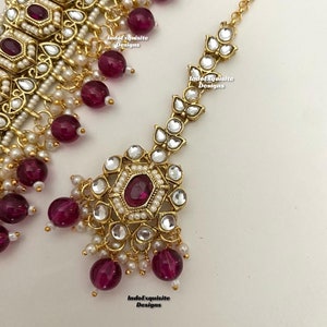 May include: A gold-tone necklace and matching maang tikka (headpiece) with a floral design. The necklace features a row of round, white pearls and burgundy-colored beads. The maang tikka has a similar design with a larger, central burgundy-colored stone.