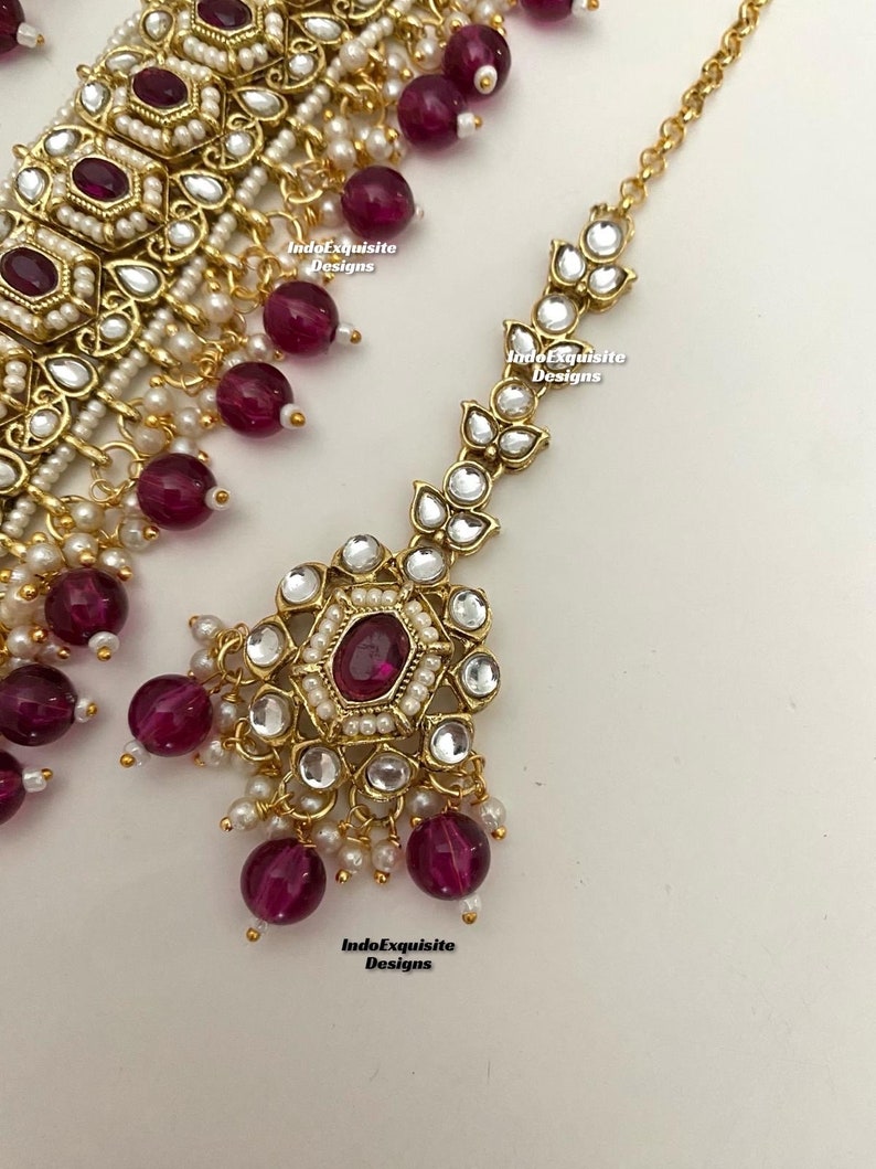 May include: A gold-tone necklace and matching maang tikka (headpiece) with a floral design. The necklace features a row of round, white pearls and burgundy-colored beads. The maang tikka has a similar design with a larger, central burgundy-colored stone.