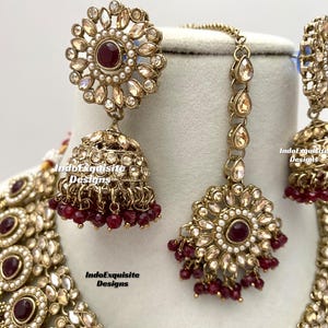 May include: A gold and white Indian bridal necklace set with red beads and a matching pair of earrings. The set features intricate floral designs and a large, round pendant.