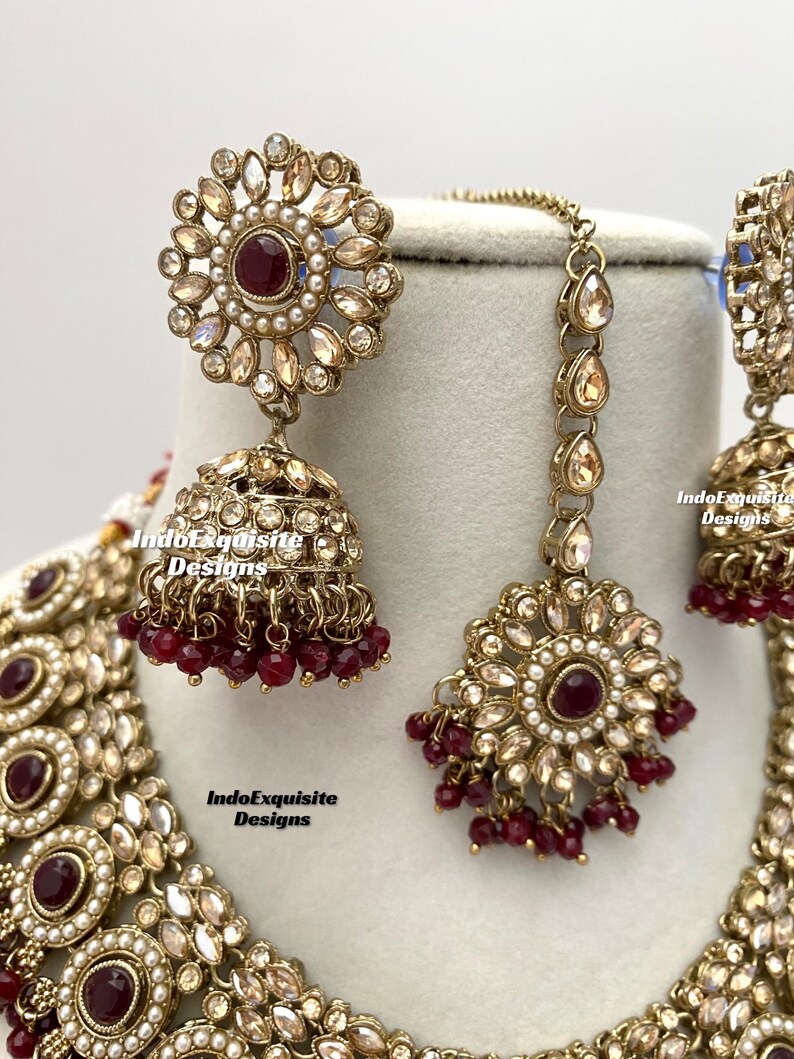 May include: A gold and white Indian bridal necklace set with red beads and a matching pair of earrings. The set features intricate floral designs and a large, round pendant.