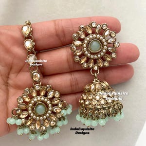 May include: A pair of gold-tone earrings with a round, floral design and a dangling teardrop-shaped charm. The earrings are adorned with clear and white stones, and the teardrop charm is accented with a light blue stone. The earrings are hanging from a hand.