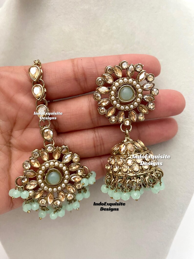 May include: A pair of gold-tone earrings with a round, floral design and a dangling teardrop-shaped charm. The earrings are adorned with clear and white stones, and the teardrop charm is accented with a light blue stone. The earrings are hanging from a hand.