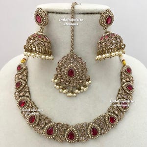 May include: An antique gold-toned jewelry set featuring a necklace, earrings, and a headpiece. The set is adorned with red teardrop-shaped stones and small white pearls. The earrings are bell-shaped, and the headpiece has a central pendant. The brand name "IndoExquisite Designs" is visible.
