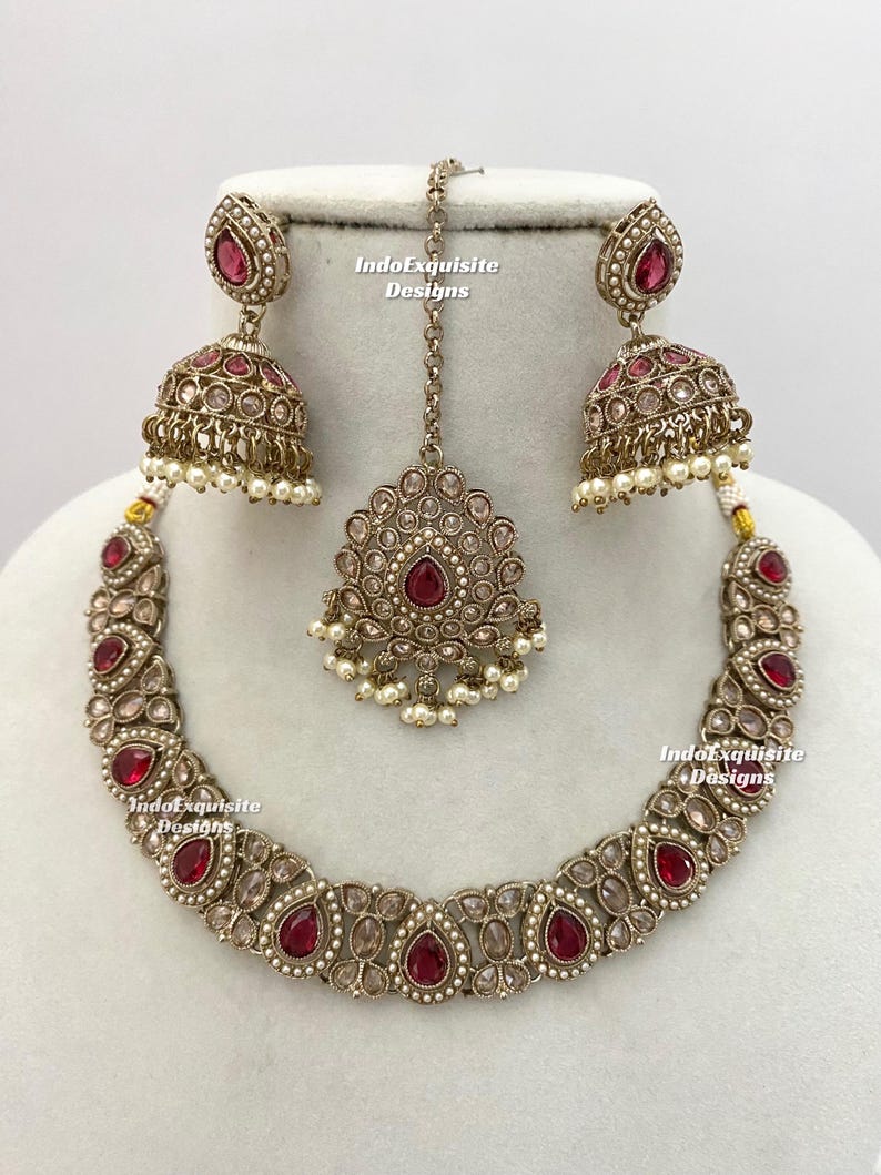 May include: An antique gold-toned jewelry set featuring a necklace, earrings, and a headpiece. The set is adorned with red teardrop-shaped stones and small white pearls. The earrings are bell-shaped, and the headpiece has a central pendant. The brand name "IndoExquisite Designs" is visible.
