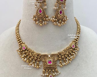 Puede incluir: Un conjunto de collar y pendientes en tono dorado con piedras rosas cuadradas y en forma de lágrima, filigrana de metal intrincada y pequeñas cuentas blancas. Las piezas de joyería llevan el texto IndoExquisite Designs.