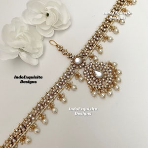 May include: A gold-tone headpiece with white pearls and clear rhinestones. The headpiece has a central design with a large clear rhinestone and smaller rhinestones arranged in a circular pattern. The headpiece is decorated with small white pearls and has a dangling design with a large clear rhinestone and smaller rhinestones. The headpiece is on a white background. IndoExquisite Designs is written on the image.