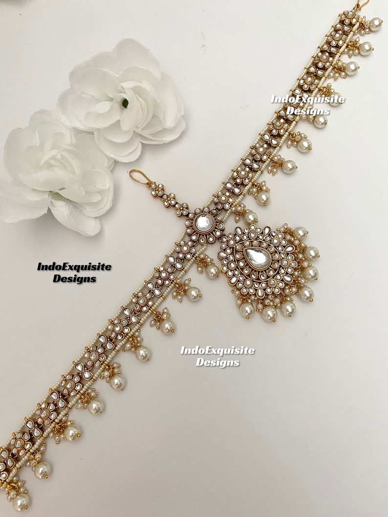 May include: A gold-tone headpiece with white pearls and clear rhinestones. The headpiece has a central design with a large clear rhinestone and smaller rhinestones arranged in a circular pattern. The headpiece is decorated with small white pearls and has a dangling design with a large clear rhinestone and smaller rhinestones. The headpiece is on a white background. IndoExquisite Designs is written on the image.
