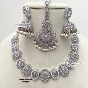 Op de afbeelding: Een zilveren ketting, oorbellen en een maang tikka-set met witte stenen en parel accenten. De ketting is een choker-stijl met ronde hangers. De oorbellen zijn hangend met een belvormige hanger. De maang tikka is een traditioneel Indiaas hoofdtooi met een hanger.