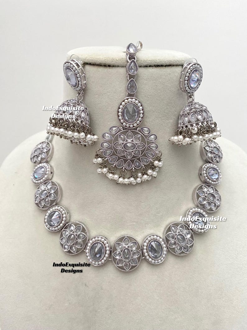 Op de afbeelding: Een zilveren ketting, oorbellen en een maang tikka-set met witte stenen en parel accenten. De ketting is een choker-stijl met ronde hangers. De oorbellen zijn hangend met een belvormige hanger. De maang tikka is een traditioneel Indiaas hoofdtooi met een hanger.