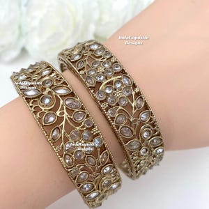 Peut inclure: Deux bracelets dorés avec des découpes florales et de feuilles complexes, ornés de pierres précieuses claires et étincelantes. Les bracelets sont présentés sur un poignet, mettant en valeur leur design orné. Le texte "IndoExquisite Designs" est visible.