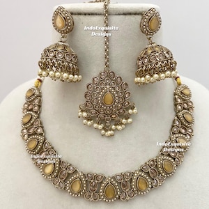 May include: A gold-toned jewelry set featuring a necklace, earrings, and a headpiece. The set includes teardrop-shaped and round gemstones, with small white pearls. The earrings are bell-shaped. The text "IndoExquisite Designs" is visible.