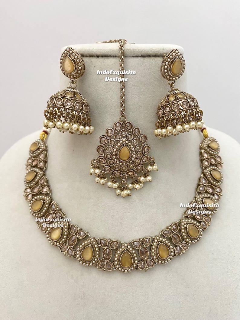 May include: A gold-toned jewelry set featuring a necklace, earrings, and a headpiece. The set includes teardrop-shaped and round gemstones, with small white pearls. The earrings are bell-shaped. The text "IndoExquisite Designs" is visible.