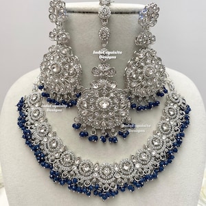 May include: A silver-coloured jewellery set comprising a necklace, earrings, and a headpiece. The set is decorated with clear crystals and dark blue beads. The necklace and earrings have a circular design, and the headpiece is a smaller, matching pendant.