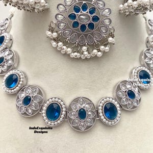 Op de afbeelding: Een zilveren ketting met blauwe en witte stenen. De ketting bestaat uit ovale hangers met een bloemmotief. De ketting is versierd met kleine witte parels. De ketting wordt geleverd met een bijpassend paar oorbellen.