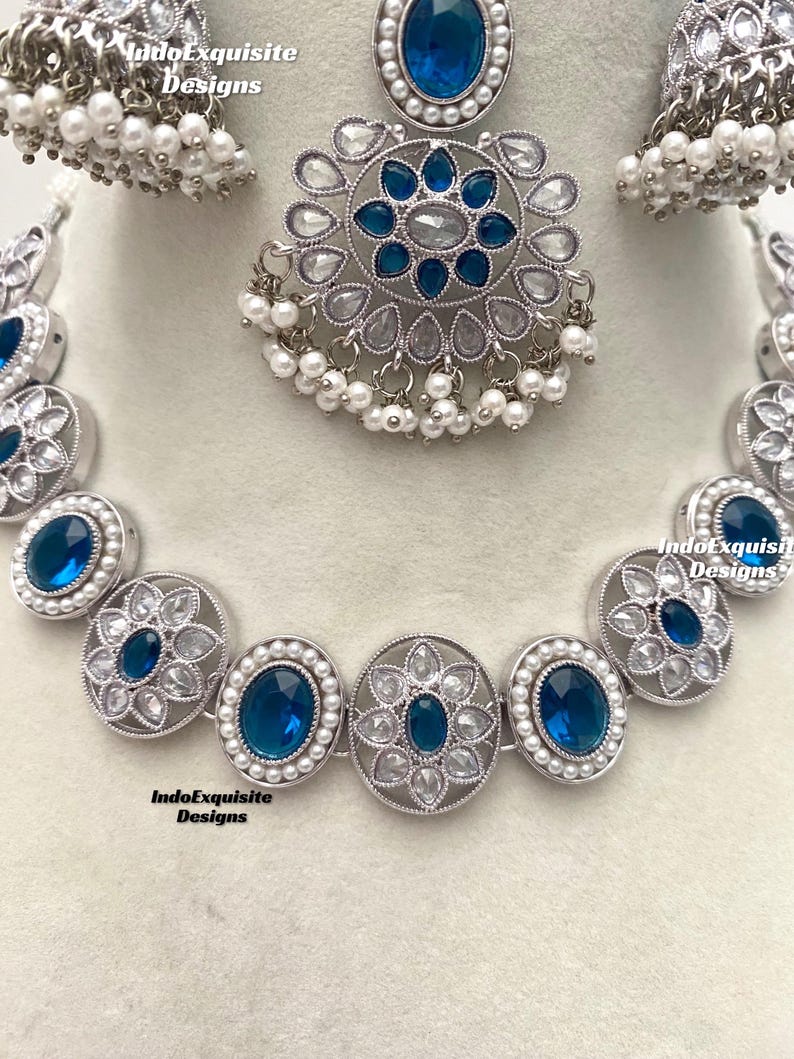 Op de afbeelding: Een zilveren ketting met blauwe en witte stenen. De ketting bestaat uit ovale hangers met een bloemmotief. De ketting is versierd met kleine witte parels. De ketting wordt geleverd met een bijpassend paar oorbellen.