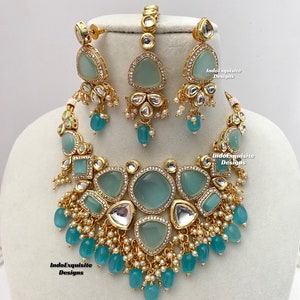 May include: A gold necklace set featuring a light blue and white stone design. The necklace has a large pendant in a teardrop shape and smaller pendants with round and square shapes. The earrings are teardrop shaped with a light blue stone. The set is accented with white pearls and gold beads.