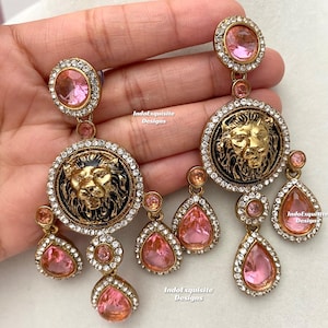 May include: A pair of gold-tone earrings with pink crystal accents. Each earring features a round pendant with a lion's face and a row of smaller pink crystal drops.