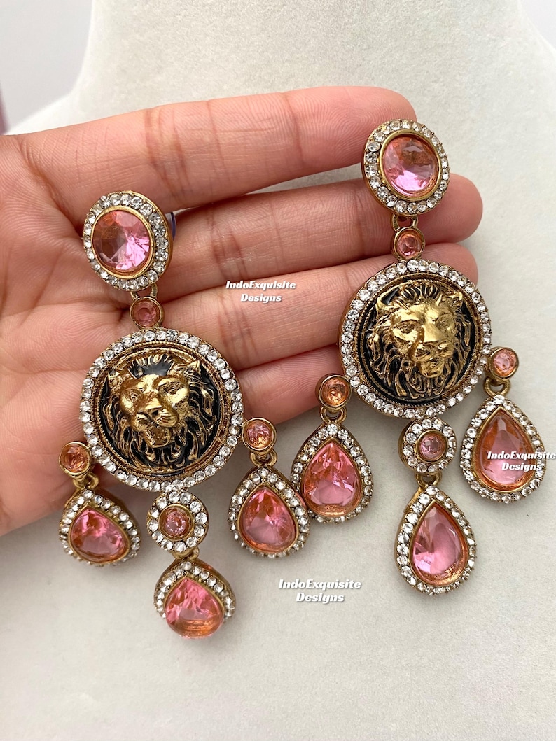 May include: A pair of gold-tone earrings with pink crystal accents. Each earring features a round pendant with a lion's face and a row of smaller pink crystal drops.