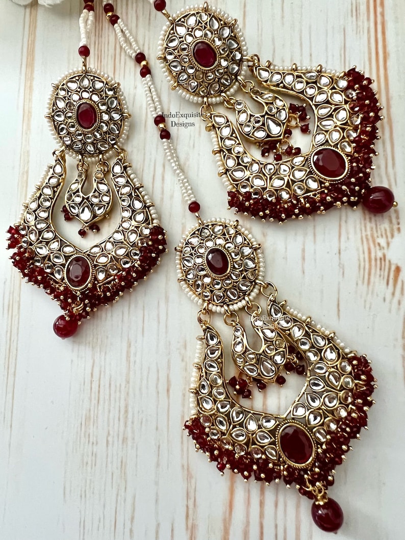 May include: A pair of gold-tone earrings with red and clear stones. The earrings are made of metal and have a traditional Indian design. The earrings are hanging from a string of white beads.