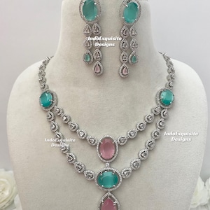 May include: A silver necklace and earring set featuring teal and pink gemstones. The necklace has two layers with oval and teardrop-shaped stones. The earrings have a cascading design with multiple stones. The brand name "IndoExquisite Designs" is visible.
