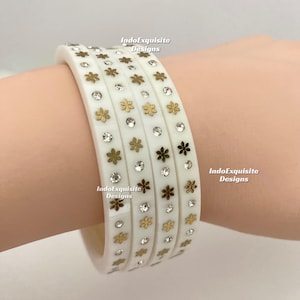 May include: White bangles featuring gold flower and clear crystal embellishments. The bangles are decorated with small gold flowers and clear crystals. The text "IndoExquisite Designs" is visible on the bangles.