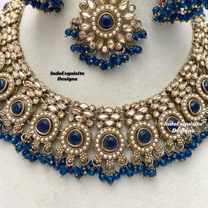 May include: A gold necklace with blue and white beads, a matching pair of earrings, and a matching maang tikka. The necklace features a central design of five round pendants with blue stones surrounded by white pearls. The earrings and maang tikka are also adorned with blue stones and white pearls. The text "IndoExquisite Designs" appears three times on the image.