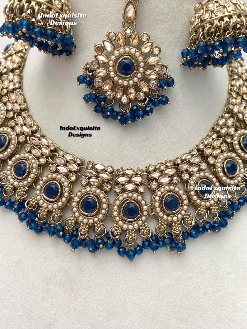 May include: A gold necklace with blue and white beads, a matching pair of earrings, and a matching maang tikka. The necklace features a central design of five round pendants with blue stones surrounded by white pearls. The earrings and maang tikka are also adorned with blue stones and white pearls. The text "IndoExquisite Designs" appears three times on the image.