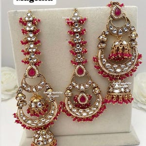 Puede incluir: Un conjunto de joyer&iacute;a en tono dorado con tres piezas que incluyen piedras magenta en forma de l&aacute;grima, piedras transparentes incrustadas y dijes colgantes en forma de campana. Las piezas tienen peque&ntilde;as cuentas magenta. El texto "Magenta" es visible.