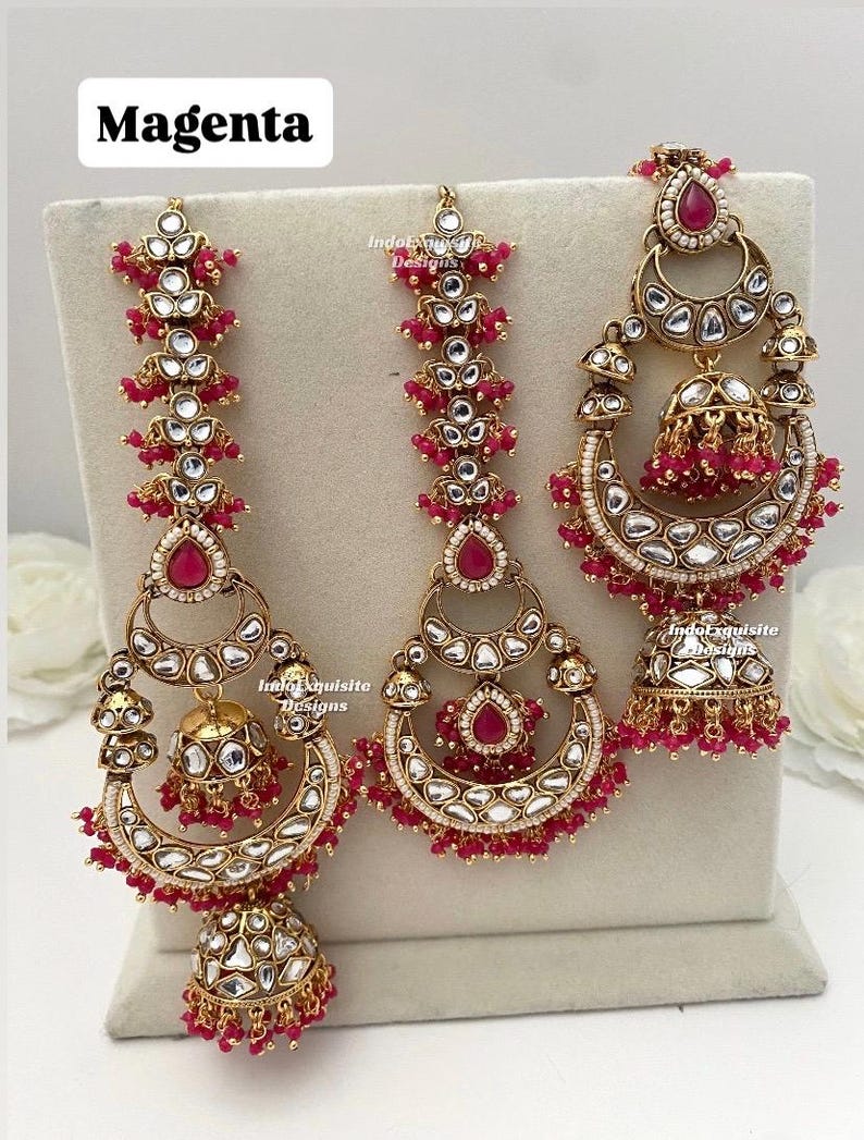Puede incluir: Un conjunto de joyer&iacute;a en tono dorado con tres piezas que incluyen piedras magenta en forma de l&aacute;grima, piedras transparentes incrustadas y dijes colgantes en forma de campana. Las piezas tienen peque&ntilde;as cuentas magenta. El texto "Magenta" es visible.