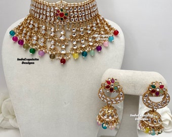 May include: A gold necklace set with a multi-layered choker featuring white stones and colorful beads. The set includes matching earrings with a crescent moon design and dangling colorful beads.