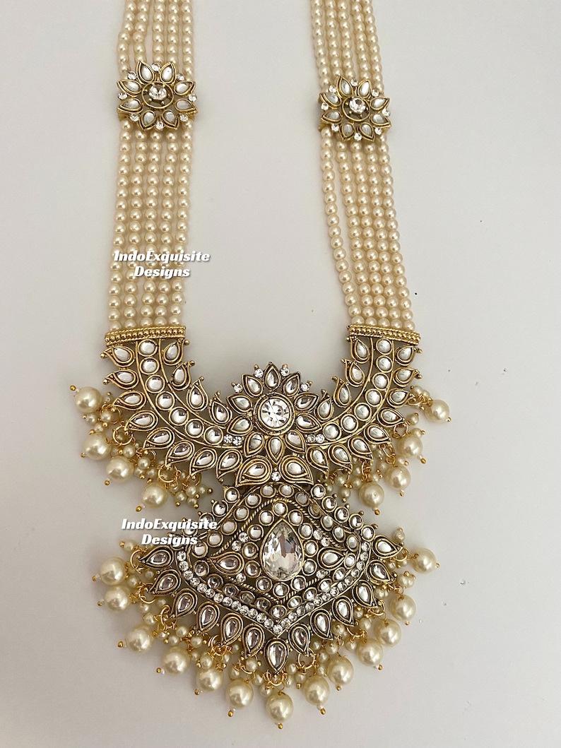 May include: An ornate necklace featuring multiple strands of faux pearls. The necklace has gold-toned metal accents with clear, sparkling stones and dangling pearl drops. The design includes floral and geometric shapes, creating an elegant and detailed piece of jewelry.