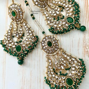 May include: A pair of gold earrings with green and white stones, and a matching necklace with green beads. The earrings are in the shape of a crescent moon and have a large, round stone in the center. The necklace is a simple chain with green beads.