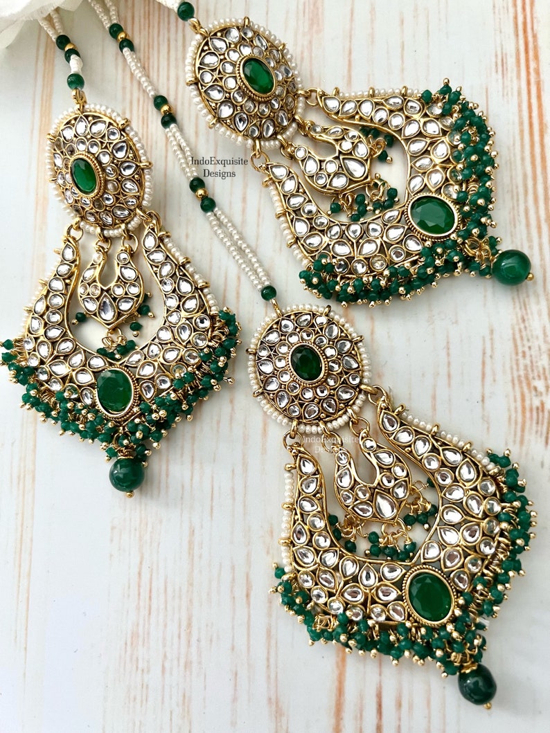 May include: A pair of gold earrings with green and white stones, and a matching necklace with green beads. The earrings are in the shape of a crescent moon and have a large, round stone in the center. The necklace is a simple chain with green beads.