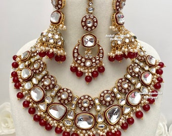 May include: A maroon and gold Indian bridal necklace set with a large, clear, faceted stone pendant. The set includes earrings and a maang tikka, all adorned with red beads and white pearls.