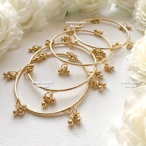 Peut inclure: Bracelets dorés avec des breloques perlées pendantes. Les bracelets sont superposés, avec le texte "IndoExquisite Designs" visible. Le fond présente des fleurs blanches. Bijoux de style traditionnel.