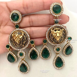 May include: Gold tone earrings with a lion head design and green and clear crystal accents. The earrings are dangling and have a teardrop shape.
