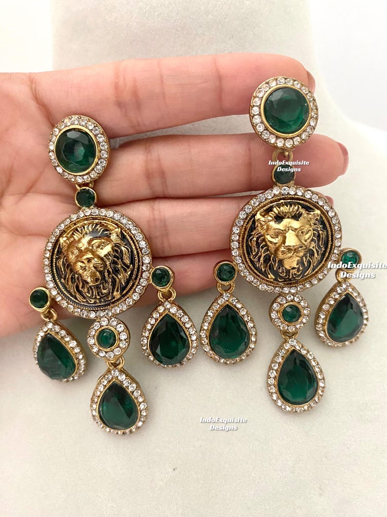 May include: Gold tone earrings with a lion head design and green and clear crystal accents. The earrings are dangling and have a teardrop shape.