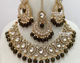 Puede incluir: Un conjunto de joyas dorado que incluye un collar, pendientes y una diadema. El conjunto está adornado con piedras transparentes de corte cuadrado, pequeñas perlas blancas y cuentas de color verde oliva. El texto "IndoExquisite Designs" es visible.