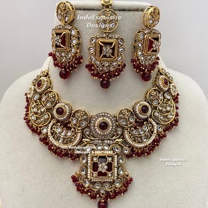 Puede incluir: Un conjunto de joyería en tono dorado que incluye un collar, dos pendientes y un maang tikka. Cada pieza está adornada con piedras cuadradas de color rojo intenso, perlas blancas y metalistería floral. El texto IndoExquisite Designs es visible.