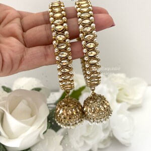 May include: A pair of gold bangles with white pearl accents and dangling gold earrings with white pearl accents. The bangles and earrings are decorated with clear stones.
